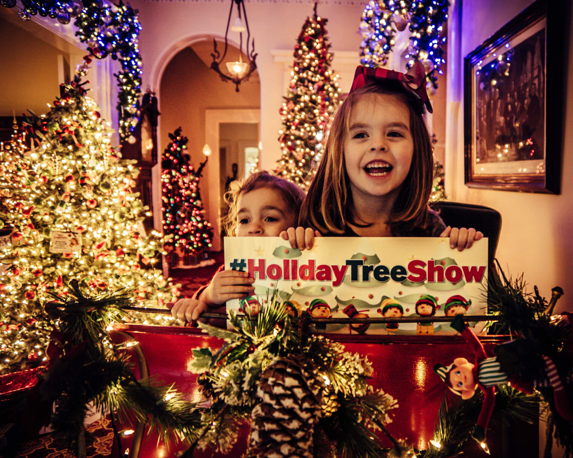 Two young girls hold a sign that says Holiday Tree Show while sitting in a sleigh in a room decorated for the holidays
